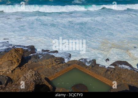 A natural rock pool filled with calm water sits on a rugged coastline, surrounded by crashing waves and turquoise ocean under bright sunlight in Praia Stock Photo