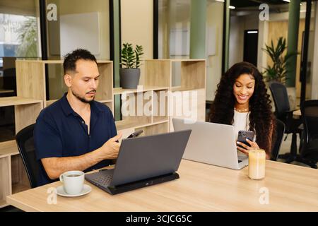 A diverse team of professionals working in a modern coworking space. Two colleagues, both focused on their laptops and smartphones, are engaging in pr Stock Photo
