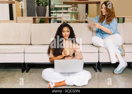Two diverse coworkers enjoy a productive and relaxed day at the office. One works on a laptop while sipping coffee, and the other engages with a table Stock Photo