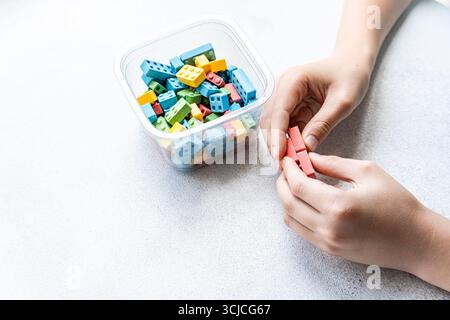 Cropped unrecognizable hands arrange colorful block candies. A container holds sweet blocks in blue, yellow, green, and red, set on a white background Stock Photo