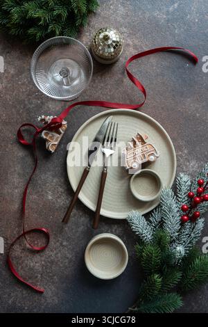 A cozy Christmas table setting featuring a gingerbread cookie on a decorative plate, festive red ribbon, evergreen branches, and a disco ball ornament Stock Photo