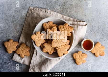 Top view of maple leaf shaped cookies in a white bowl on a gray surface. The cookies rest on a beige cloth, accompanied by a small cup of maple syrup. Stock Photo