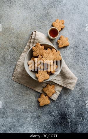Top view of maple leaf shaped cookies in a white bowl on a beige linen napkin. A small bowl of maple syrup is nearby, set against a textured gray back Stock Photo