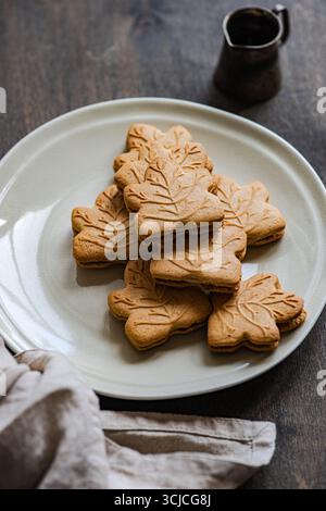 From above, a stack of maple leaf shaped cookies rests on a beige plate, set against a dark wooden background. The cookies’ intricate design adds a ho Stock Photo