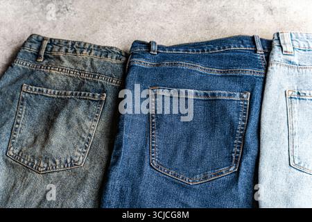 Three pairs of jeans in different washes lined up side by side on a textured surface. Each pair showcases the distinct shades and styles in casual den Stock Photo