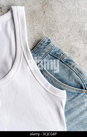 A simple composition of a white ribbed tank top placed next to a pair of classic blue jeans on a textured gray surface, epitomizing casual style in ev Stock Photo