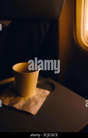 A paper cup rests on a napkin atop an airplane tray table, illuminated by soft, warm sunset light from the adjacent window, capturing a peaceful in-fl Stock Photo