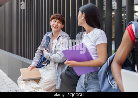 A caucasian boy and an asian girl, university students converse happily outdoors, showcasing friendship and cultural diversity. They hold notebooks an Stock Photo