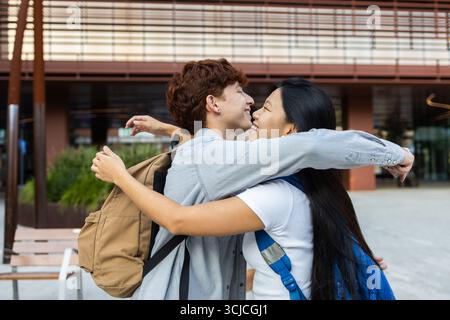 Two happy multiethnic university students hug joyfully outside a campus building, celebrating friendship and diversity. Both wear backpacks, signifyin Stock Photo