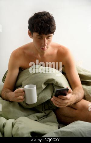 A young man sits in bed wrapped in a green comforter, holding a white mug and looking at his smartphone. The scene suggests a relaxed morning ritual a Stock Photo