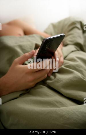 A young man is lying in bed, casually using his smartphone. The scene captures a relaxed moment, highlighting the contemporary lifestyle of digital co Stock Photo