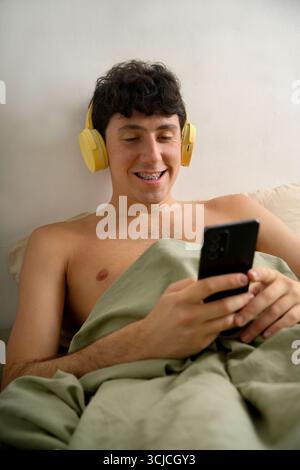 A young man with headphones relaxes in bed under a blanket, smiling while looking at his smartphone, enjoying a comfortable morning in a warm and cozy Stock Photo