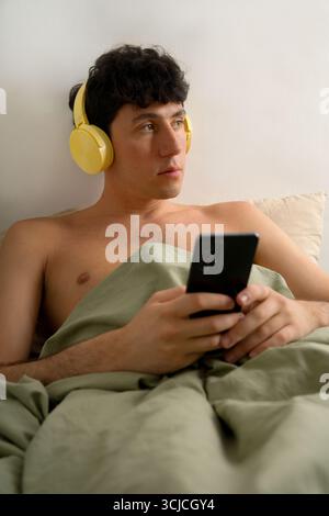A young man lies in bed with a relaxed expression, wearing yellow headphones and holding a smartphone. The setting suggests a lazy morning focused on Stock Photo