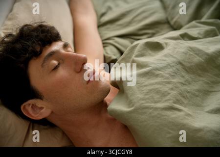 A young man lies peacefully in bed, wrapped in a soft green blanket, with a calm and serene expression, representing a quiet morning moment of rest Stock Photo