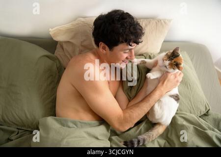 A young man relaxes in bed, lovingly playing with his cat. The cozy setting suggests a peaceful morning routine filled with affection and companionshi Stock Photo