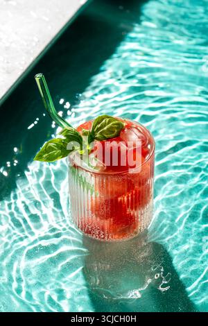 A refreshing glass of watermelon beverage with ice, garnished with fresh basil leaves, sits in the shimmering turquoise pool water, evoking a perfect Stock Photo