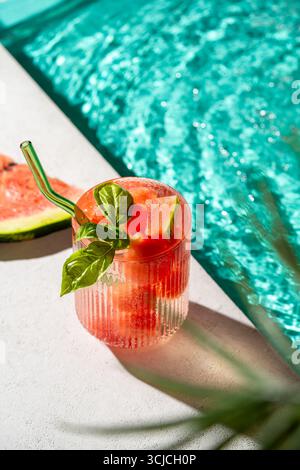 A refreshing watermelon drink in a glass with a green straw, garnished with fresh mint leaves, set on a poolside table. Sunlight reflects off the turq Stock Photo