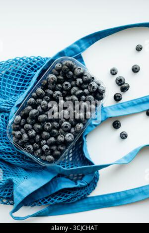 Fresh blueberries in a transparent container placed on a blue eco-friendly reusable mesh bag, showcasing sustainable practices in food storage and sho Stock Photo