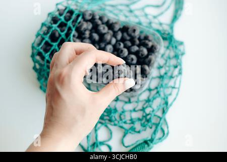 A close-up image of a hand holding a fresh blueberry over a container and an eco-friendly green mesh bag, highlighting sustainable and natural lifesty Stock Photo
