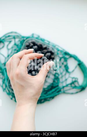 A hand delicately holds a ripe blueberry above a pile of berries inside an eco-friendly, reusable green mesh bag, highlighting sustainable and healthy Stock Photo