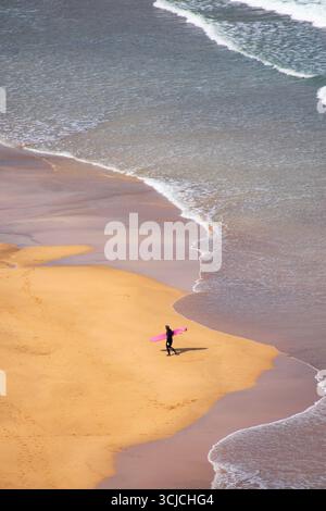 A lone surfer with a vibrant pink surfboard walks along the sandy shore of Zarautz beach. The waves gently lap at the coastline, capturing a moment of Stock Photo
