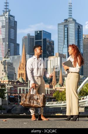 A diverse group of young professionals engages in a lively outdoor discussion against the vibrant cityscape of Melbourne, embodying modern urban busin Stock Photo