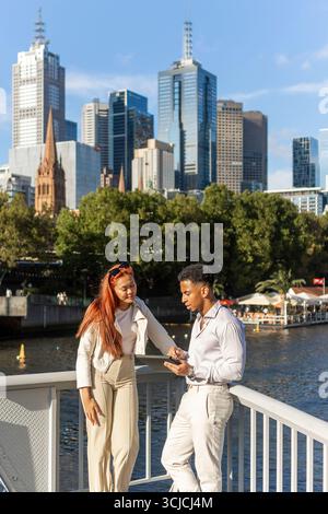 Multicultural professionals having a business discussion on Melbourne's Southbank promenade, with the city skyline as their backdrop, utilizing a digi Stock Photo