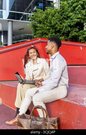 Multicultural professionals engaged in a casual outdoor business meeting in Melbourne. A woman and man discussing work using a digital device, showcas Stock Photo