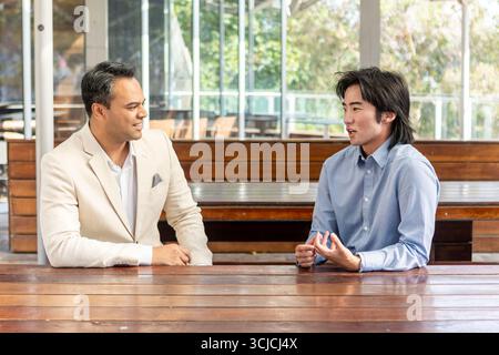 Two professional men engaged in a business discussion, seated at a wooden table in Melbourne. The atmosphere is casual yet focused, emphasizing urban Stock Photo