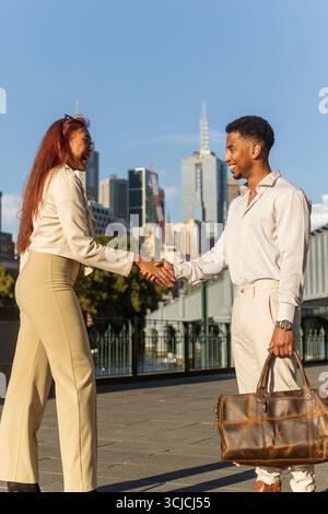 A man and a woman, dressed in business casual attire, shake hands outdoors in front of the Melbourne city skyline, exemplifying multicultural business Stock Photo