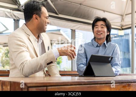 Two professional men in Melbourne engage in a lively business conversation outdoors, enjoying the sunny day. One holds a coffee, while the other has a Stock Photo
