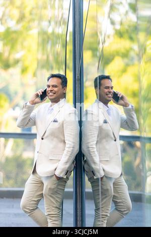 A professional man in a beige suit makes a cheerful phone call while leaning against a reflective glass in Melbourne, with greenery and a modern citys Stock Photo