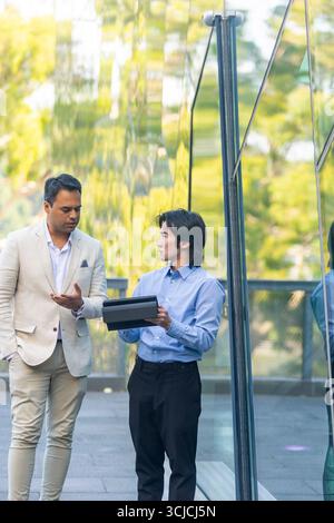 Two men engage in a professional conversation against a modern glass building in Melbourne. They appear focused and thoughtful, reflecting urban busin Stock Photo