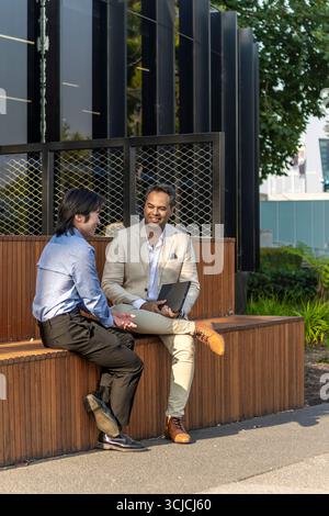 Two professional men engage in a business conversation outdoors in Melbourne. Sitting on a bench, they are focused on discussion, reflecting an urban Stock Photo