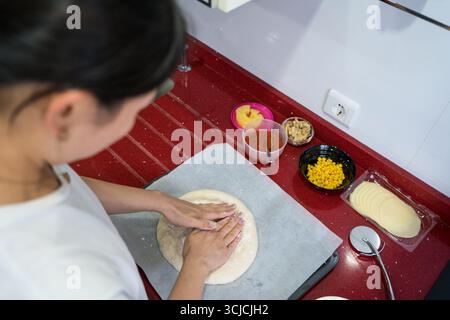 A woman kneads dough on a red countertop, surrounded by bowls of toppings like cheese, corn, and sauce, illustrating the joy of making pizza at home Stock Photo