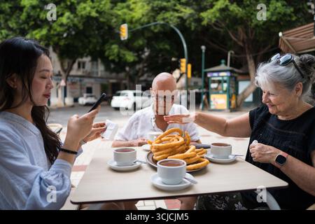 A multiethnic family enjoys a traditional Spanish snack of churros with hot chocolate at an outdoor cafe. Three people are seated at a wooden table, s Stock Photo