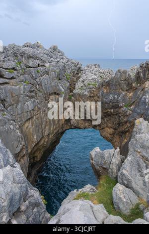 Spectacular view of Acantilados del Infierno with dramatic cliffs and a natural stone arch. A lightning bolt strikes over the ocean, highlighting the Stock Photo