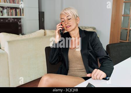 Professional woman in a black blazer working from home, sitting at a table with a laptop, talking on the phone, navigating the challenges of remote wo Stock Photo