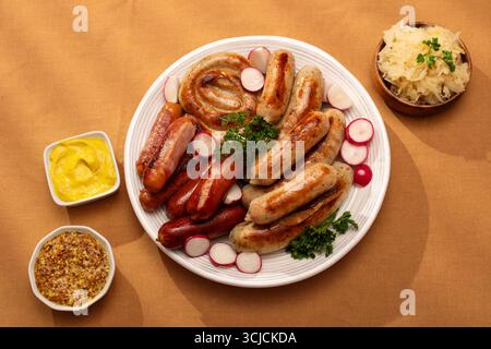 Top view of a variety of grilled German sausages both Bratwurst, Frankfurters on a white plate garnished with radish slices and parsley. Served with m Stock Photo