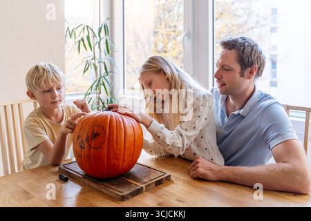 A father and his two children are carving a Halloween pumpkin together at the dining table, enjoying a fun and creative family activity during the aut Stock Photo