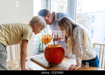 A family gathers in their cozy home to carve a Halloween pumpkin. A father and two children work together, scooping out the seeds, enjoying creative a Stock Photo