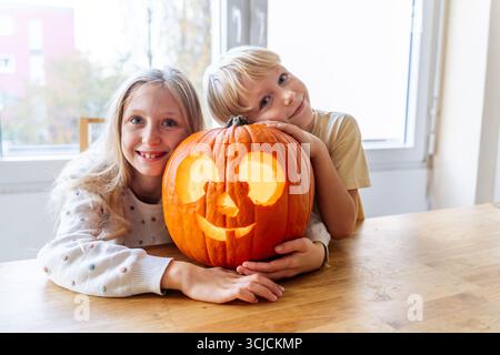 Two siblings happily pose with their carved Halloween pumpkin, showcasing a delightful autumn tradition. The children are dressed warmly, embodying th Stock Photo