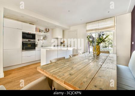 Bright modern kitchen interior featuring sleek white cabinets and a rustic wooden dining table. Natural light fills the space, highlighting elegant de Stock Photo