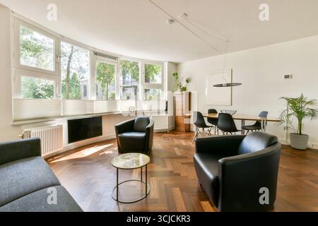 A bright living room featuring contemporary minimalist design with large windows, black leather furniture, wood flooring, and a dining area, creating Stock Photo
