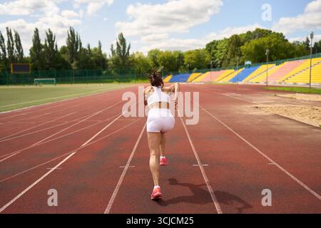 An athlete in a white sports outfit is captured mid-sprint on an outdoor track, demonstrating determination and speed. The empty stadium seats and gre Stock Photo