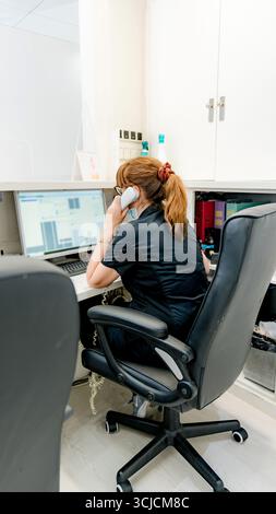 A dental office receptionist is multitasking by taking a phone call while working on a computer. The modern, organized workspace emphasizes profession Stock Photo