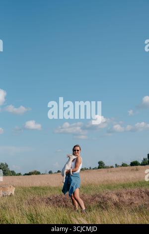 A smiling woman wearing sunglasses holds a goat in a sunlit farm field. She stands on green grass under a blue sky, capturing a serene moment of rural Stock Photo