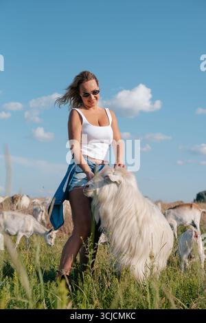 A smiling woman wearing sunglasses stands in a sunlit field surrounded by goats, enjoying the beautiful farm environment. Clear blue sky with clouds h Stock Photo