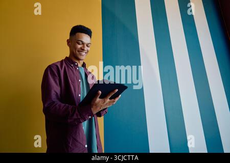 Smiling young businessman in smart casual using tablet while standing against colorful wall in office Stock Photo