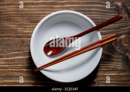 A pair of wooden chopsticks and spoon lying across an empty white soup bowl on wooden table, close up, top view Stock Photo